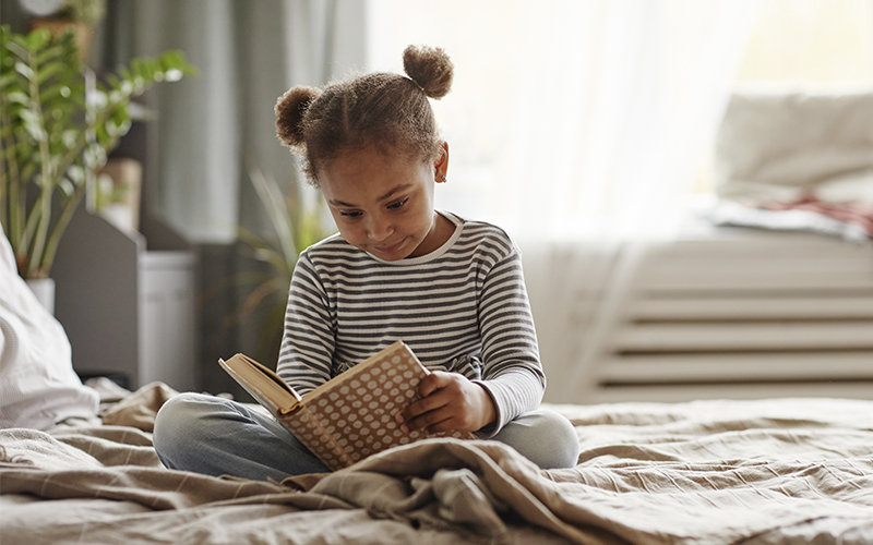Fotografia de uma menina sentada na cama lendo um livro 