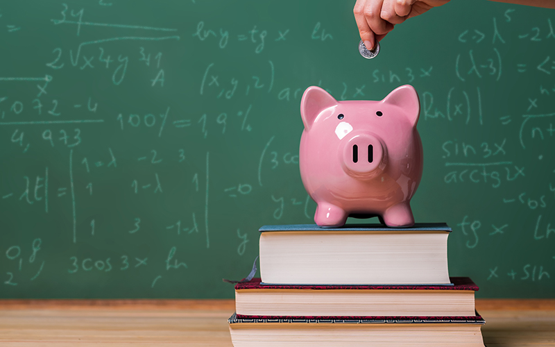 Foto de um porquinho de dinheiro com um quadro de fundo, remetendo a sala de aula